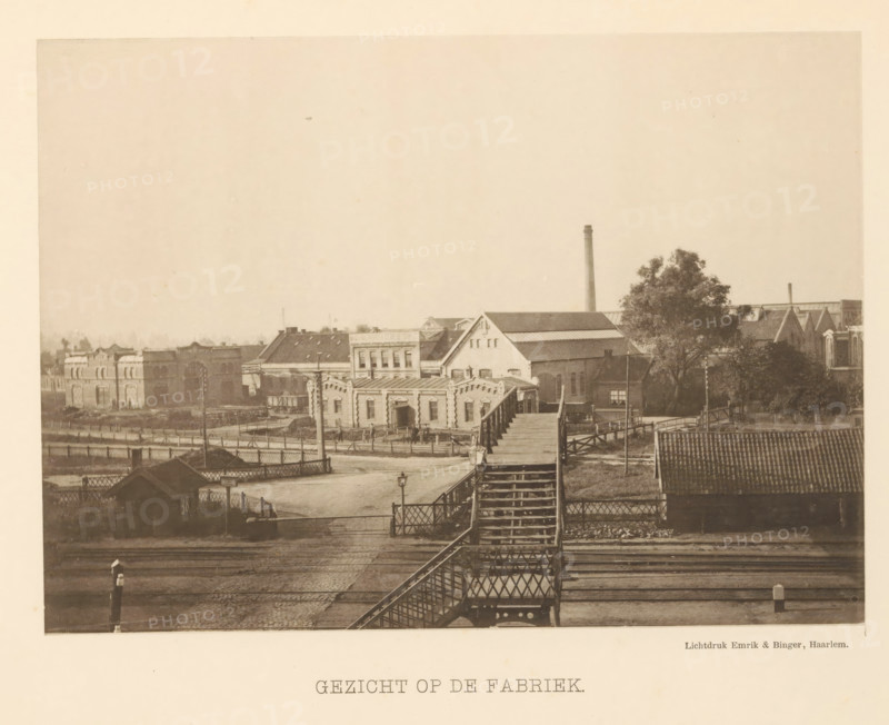 The machine factory of Gebroeders Stork & Co., in Hengelo, commemorative publication on the occasion of the twenty-fifth anniversary on September 4, 1893., book, printer (person): Mouton & Co., The Hague, 1894, printer (person), textile materials, pasteboard, paper, linen (material), collotype, decorated paper, height 335 mm x width 265 mm x thickness 20 mm, A sepia-toned photograph showing a row of industrial and residential buildings behind a wooden pedestrian bridge that spans over railway tracks, the bridge featuring lattice side railings and a long flight of stairs leading down to a fenced area; the foreground includes cobbled ground, a small lamp post, and a posts beside the tracks, while the background shows varied rooftops, a tall smokestack, a large tree near a cluster of buildings, and a wooden structure with a pitched roof at the right edge.