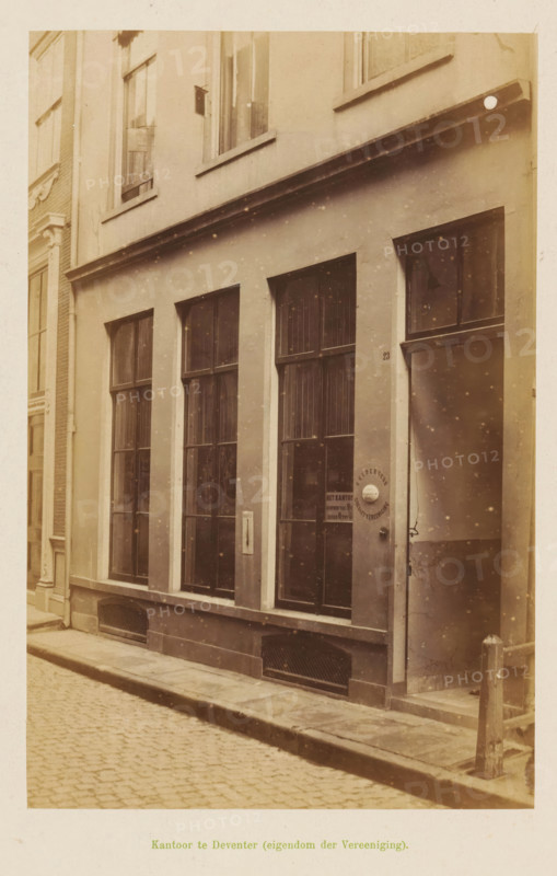 View of the office of the Geldersche Crediet-Vereeniging in Deventer, Office in Deventer (property of the Association), photograph, page, Deventer, c. 1891 - c. 1892, photographer, paper, albumen print, height 193 mm x width 126 mm, façade (of house or building), A sepia-toned street scene showing the facade of a narrow building with tall, divided windows set in a smooth plastered wall, a recessed doorway with a round door handle plate, a column-like wall edge at the corner, a raised curb and cobblestone pavement in front, narrow rectangular ventilation grilles at the base of the facade, a small metal post near the entrance, upper floor windows with sills, and subtle speckled aging marks across the photograph.