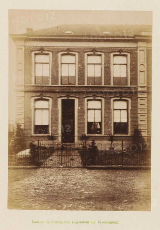 View of the office of the Geldersche Crediet-Vereeniging in Doetinchem, Office in Doetinchem (property of the Association), photograph, page, Doetinchem, c. 1891 - c. 1892, photographer, paper, albumen print, height 155 mm x width 111 mm, façade (of house or building), A two-story brick building facade with a central doorway flanked by tall rectangular windows on each side and matching windows above, decorative stone trim around the windows and along the building corners, a low iron fence and a paved path leading to the entrance, small plants and shrubs in front of the building, a tiled roof with roof windows partially visible, and a cobblestone street in the foreground.