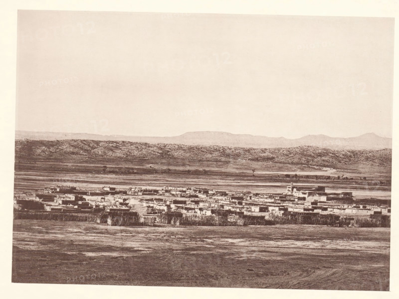 Pueblo of Santo Domingo, page, photomechanical print, number: 'Fig. 22.', photographer: John Karl Hillers, New Mexico, cliché maker: Heliotype Printing Company, Boston (Massachusetts), c. 1876 - in or before 1881, North America, Massachusetts, paper, collotype, photoprint, height 160 mm x width 221 mm, city-view in general, 'veduta', Santo Domingo Pueblo, A wide, sepia-toned landscape showing a flat foreground with textured ground leading to a clustered settlement of low, rectangular buildings with flat roofs, some taller structures rising slightly above the rest; behind the settlement there is a narrow strip of lighter land and rows of vegetation, then rolling hills with mottled, bush-covered slopes, and a distant line of low mountains beneath a pale, empty sky.