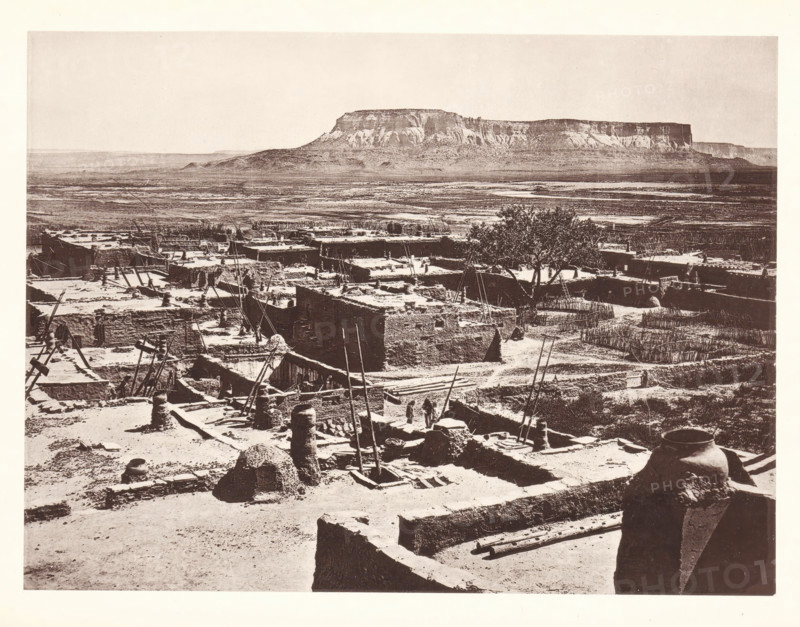 Pueblo of Zuni, page, photomechanical print, number: 'Fig. 23.', photographer: John Karl Hillers, New Mexico, cliché maker: Heliotype Printing Company, Boston (Massachusetts), c. 1876 - in or before 1881, North America, Massachusetts, paper, collotype, photoprint, height 172 mm x width 231 mm, city-view in general, 'veduta', Zuni Pueblo, A wide sepia-toned scene shows a sprawling adobe village with flat-roofed mud structures, many with scaffolding poles and ladders leaning against them, and narrow pathways winding between the buildings; in the foreground a large rounded clay jar sits on a ledge beside stacked rectangular walls and scattered wooden beams, while further back a taller tree rises above the rooftops and a distant flat-topped mesa dominates the horizon over an open plain.