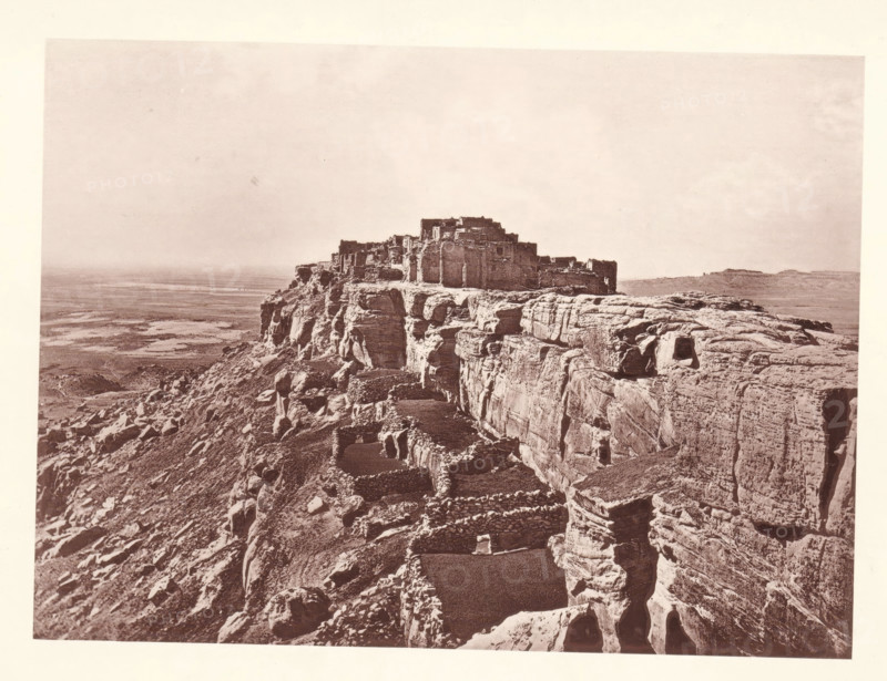 Pueblo of Wolpi, page, photomechanical print, number: 'Fig. 25.', photographer: John Karl Hillers, Arizona, cliché maker: Heliotype Printing Company, Boston (Massachusetts), c. 1876 - in or before 1881, North America, Massachusetts, paper, collotype, photoprint, height 160 mm x width 216 mm, city-view in general, 'veduta', mountains, Walpi, A sepia-toned photograph shows a rocky mesa with layered cliff faces and rough stone terraces leading up to a cluster of rectangular stone structures perched at the top; the foreground features steep, eroded rock with small recessed openings and stacked stone enclosures, while the midground reveals the fortified buildings and the background opens to a broad, flat plain under a pale sky.