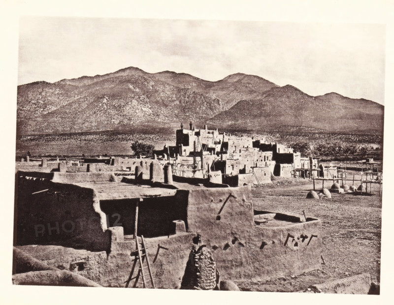 North Pueblo of Taas, page, photomechanical print, number: 'Fig. 27.', photographer: John Karl Hillers, United States of America, cliché maker: Heliotype Printing Company, Boston (Massachusetts), c. 1876 - in or before 1881, North America, Massachusetts, paper, collotype, photoprint, height 161 mm x width 218 mm, city-view in general, 'veduta', mountains, façade (of house or building), A sepia-toned scene shows a cluster of multi-level adobe buildings with flat roofs, protruding wooden beams, ladders leaning against walls, and rounded door and window openings; a foreground adobe wall with a rough opening and a leaning ladder frames the view, and a conical stone or adobe structure stands near the wall; beyond the main cluster are more adobe structures, fenced enclosures, and dome-shaped oven-like forms on the ground; a wide plain separates the settlement from a backdrop of rugged, tree-covered mountains under a pale sky.