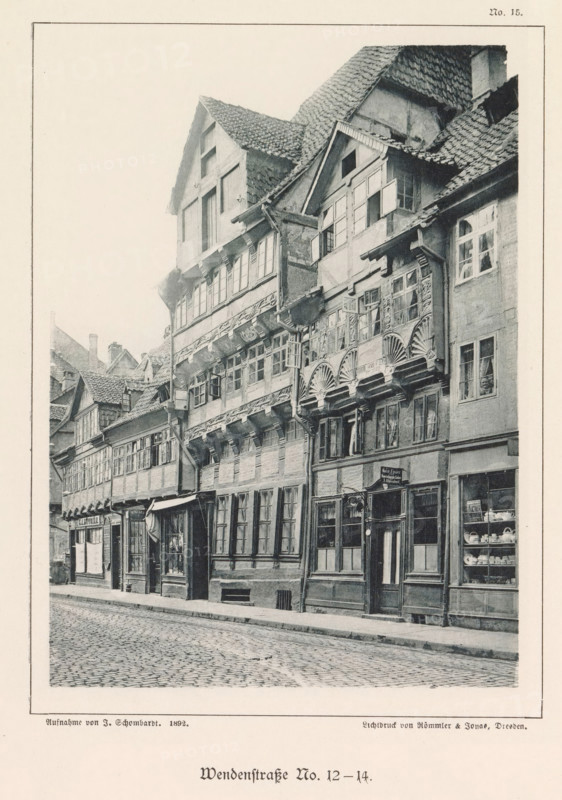 Exterior of buildings at Wendenstraße 12 and 14 in Braunschweig, Wendenstraße No. 12-14, Braunschweig's Baudenkmäler, page, photomechanical print, number: 'No. 15.', photographer: J. Schombardt, Braunschweig, cliché maker: Römmler & Jonas, Dresden, publisher: Benno und Wilhelm Danert Goeritz, 1892, J. Schombardt, Römmler & Jonas, Benno und Wilhelm Danert Goeritz, photographer, cliché maker, publisher, Lower Saxony, Saxony, paper, collotype, photoprint, height 148 mm x width 108 mm, street, facade (or house or building), A narrow cobblestone street lined with a row of tall, timber-framed buildings with overhanging upper stories and steep, tiled roofs, featuring multiple windows of various sizes and styles, some with small panes and some with shutters, ornate wooden carvings and decorative fan-shaped motifs beneath several windows, visible shopfronts at street level with display windows showing dishes and other items, a doorway with a hanging sign and a small awning, downspouts and expos...