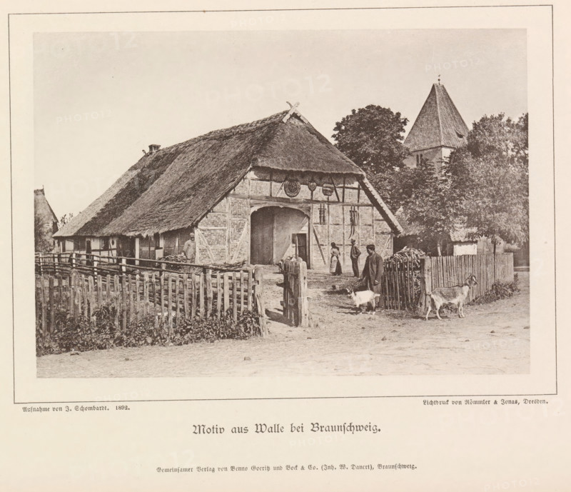 Motiv from Walle bei Braunschweig, Exterior of a farm in Walle, Braunschweig's Baudenkmäler, page, photomechanical print, number: 'No. 39.', photographer: J. Schombardt, Walle, cliché maker: Römmler & Jonas, Dresden, publisher: Benno und Wilhelm Danert Goeritz, Braunschweig, 1892, J. Schombardt, Römmler & Jonas, Benno und Wilhelm Danert Goeritz, photographer, cliché maker, publisher, Lower Saxony, Saxony, paper, collotype, photoprint, height 105 mm x width 151 mm, farm (building), facade (or house or building), A sepia-toned photograph of a rural scene showing a thatched-roof farmhouse with exposed timber framing and a large arched entrance, a wooden picket fence in front, a few people standing near the entrance and along the yard, two goats in the foreground near the gate, stacks of firewood beside the fence, leafy trees behind and to the side of the house, and a steep-roofed tower partially visible among the trees.