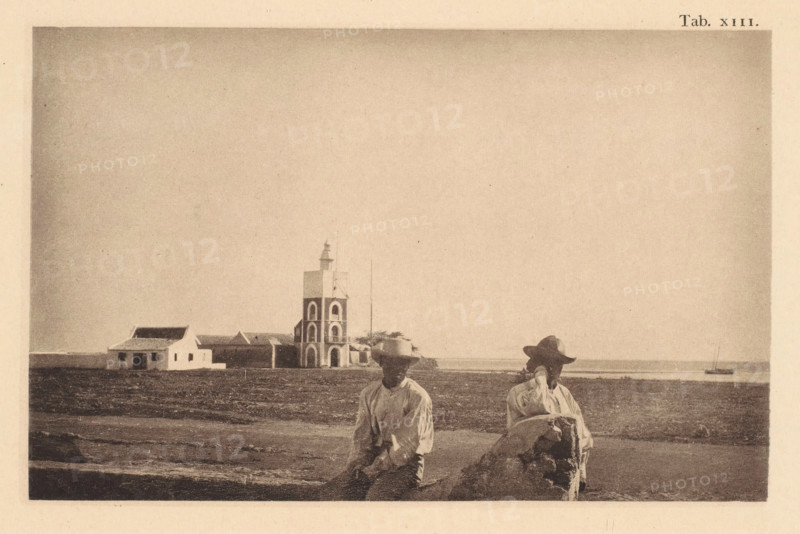 View of Fort Zoutman in Oranjestad on Aruba, page, photomechanical print, number: 'Tab. XIII.', photographer: Jacobus Rudolphus Neervoort van de Poll (possibly), Oranjestad (Aruba), cliché maker: anonymous, c. 1877 - in or before 1887, Aruba, paper, collotype, photoprint, height 105 mm x width 166 mm, Print in section at the back., fortress, Fort Zoutman, A sepia-toned coastal scene with two seated men in the foreground wearing light shirts and wide-brimmed hats, one resting an arm on a large rock. Behind them is a flat, open shoreline leading to a small complex of buildings, including a multi-level tower with arched openings and a smaller white house with a pitched roof. A calm body of water is visible at the horizon with a small boat anchored offshore. The sky above is empty and pale.