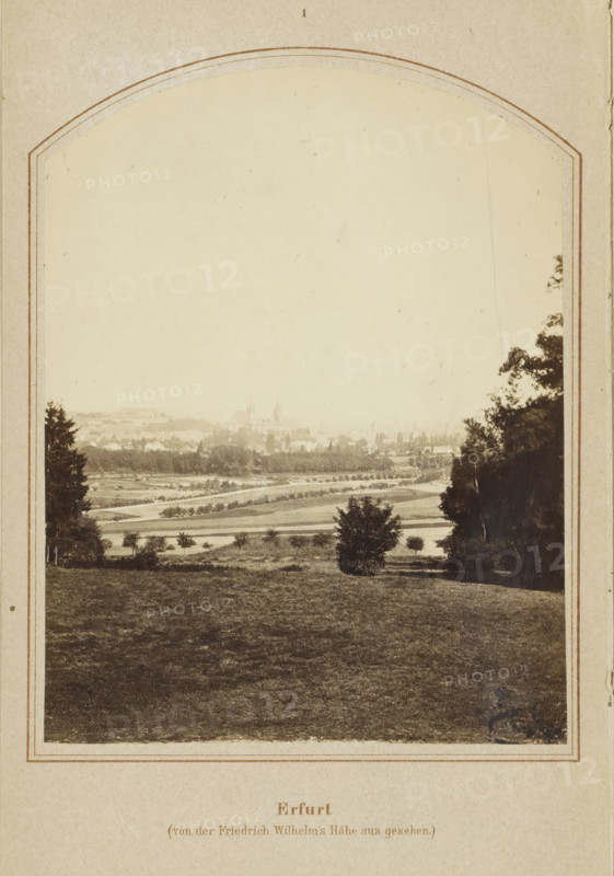 View of Erfurt, Erfurt (von der Friedrich Wilhelm's Höhe aus gesehen), photograph, page, number: '1', Erfurt (district), c. 1858 - in or before 1868, photographer, Thuringia, paper, albumen print, height 128 mm x width 98 mm, Image on frontispiece., city-view in general, 'veduta', Erfurt (city), A sepia-toned framed photograph showing a wide landscape from a slightly elevated viewpoint, with a foreground of grassy terrain that slopes gently toward a middle ground of open fields and scattered small trees and shrubs, a solitary bush near center foreground, and a winding lighter-toned path or road traversing the fields; taller trees flank the scene on the left and right edges, partially obscuring the view, while the distant background fades into a hazy skyline with faint outlines of clustered buildings and distant church spires barely visible against a pale sky.
