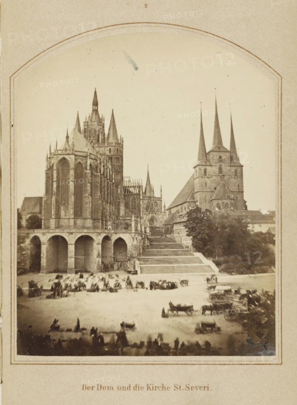 View of Erfurt Cathedral and St. Severus Church, Der Dom und die Kirche St. Severi, photograph, page, number: '2', Erfurt (city), c. 1858 - in or before 1868, photographer, Thuringia, paper, albumen print, height 128 mm x width 103 mm, Image opposite page 8., square, circus, church (exterior), façade (of house or building), Erfurt Cathedral, A sepia photograph framed by a decorative border shows a large Gothic cathedral with tall pointed spires and intricate stone facades on the left and a church with three tall slender spires on the right, connected by a raised terrace and a broad flight of steps; beneath the terrace an arcade of rounded arches is visible, and a wide open square in front of the buildings contains numerous horse-drawn carriages and groups of people, with trees and smaller buildings along the right side and a pale sky above.