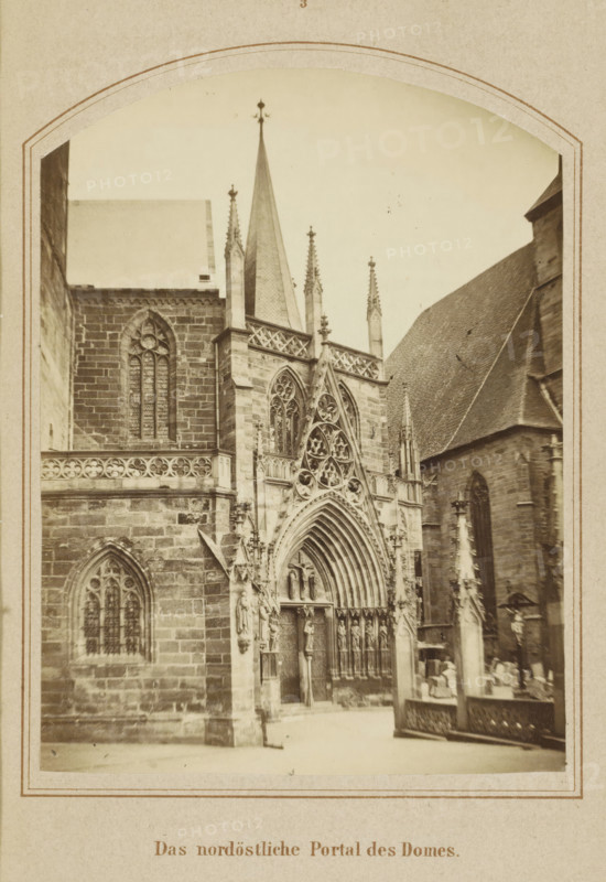The northeastern portal of the cathedral, Northeastern entrance portal of the Cathedral of Erfurt, photograph, page, number: '3', Erfurt (city), c. 1858 - in or before 1868, photographer, Thuringia, paper, albumen print, height 125 mm x width 92 mm, Image opposite page 24., façade (of house or building), parts of church exterior and annexes: portal, Cathedral of Erfurt, A sepia-toned photograph of a gothic cathedral facade framed by a decorative arched border, showing pointed spires and ornate stonework. The central portal features a large pointed arch with concentric moldings, intricate tracery, and sculpted figures in niches flanking the doorway. Tall narrow lancet windows with tracery are set into the stone walls, and a steep roof plane rises to the right. Ornamental pinnacles and finials project above the parapet and along the roofline. In the foreground a low stone balustrade and a carved column or monument with decorative elements are visible, and a few indistinct figures appear ...
