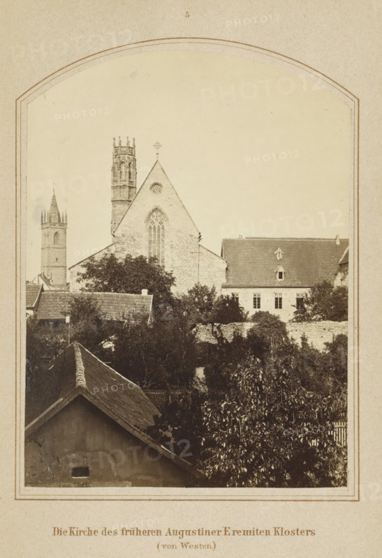 The Church of the former Augustinian Hermitage Monastery (von Westen), View of the church of the former Augustinian monastery in Erfurt, photograph, page, number: '5', Erfurt (city), c. 1858 - in or before 1868, photographer, Thuringia, paper, albumen print, height 126 mm x width 93 mm, Image opposite page 56., church (exterior), abbey, monastery, convent, Roman Catholic church, façade (of house or building), Augustinian Monastery, A sepia-toned photograph framed by an arched border shows a townscape with a large stone church as the central focus, featuring a steep gabled roof, a pointed arched window with tracery, and a circular window near the roof peak, flanked by a tall ornate tower with decorative pinnacles and a separate square tower with a spire in the background; in front of the church are clustered rooflines of smaller buildings, dense foliage from trees and bushes, and a low stone wall, while the foreground contains the peaked roof of a nearer structure and more vegetation, a...
