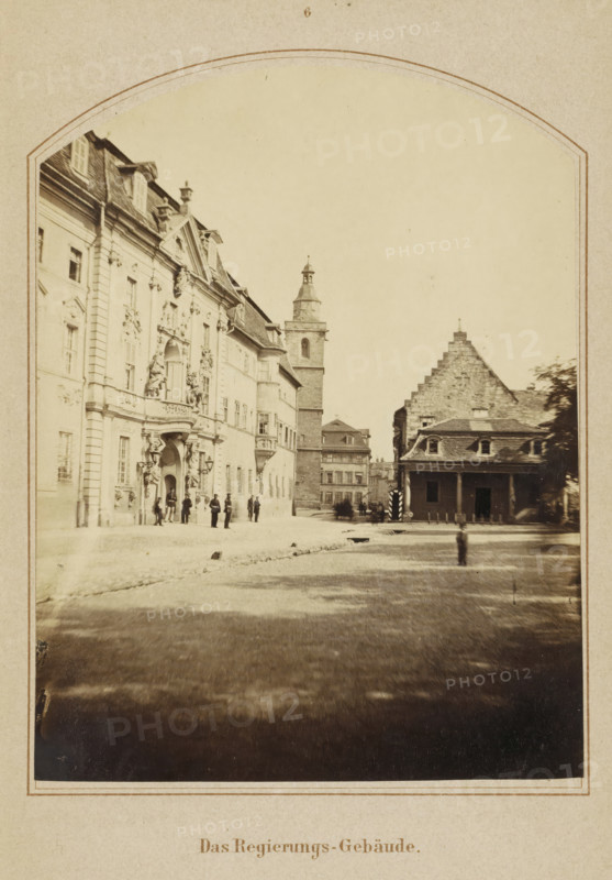 View of the Thuringian State Chancellery in Erfurt, Das Regierings-Gebäude, photograph, page, number: '6', Erfurt (city), c. 1858 - in or before 1868, photographer, Thuringia, paper, albumen print, height 130 mm x width 99 mm, Image opposite page 72., square, circus, façade (of house or building), Thüringer Staatskanzlei, A sepia-toned photograph framed by an ornate arch shows a wide street flanked by large historic buildings. On the left, a grand multi-story façade with decorative sculptures, columns, and arched doorways faces the street, and a small group of people stands near its entrance. In the center distance a tall tower rises above surrounding structures. To the right, a building with a stepped gable and a lower porch with columns sits closer to the foreground. The ground in front appears open and worn, and a single indistinct figure stands near the middle of the open area. The image has a faded, antique quality.