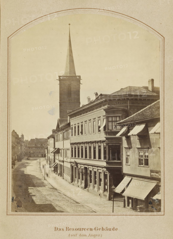 Das Resourcen-Gebäude (auf dem Anger), View of buildings along the Anger in Erfurt, photograph, page, number: '7', Erfurt (city), c. 1858 - in or before 1868, photographer, Thuringia, paper, albumen print, height 131 mm x width 99 mm, Image opposite page 88., street, square, circus, façade (of house or building), A sepia-toned street scene framed by a rounded arch shows a row of multi-story buildings lining a cobbled road, with a tall church tower and spire rising behind the structures. The foremost building on the right has a decorative cornice, a bay window, and awnings over ground-floor windows and entrances, while the adjacent façades display regularly spaced windows and ornamental details. A narrow sidewalk runs beside the buildings, and a lamppost stands near the curb. Further down the street smaller buildings and rooftops recede into the distance, and faint human figures are visible along the sidewalk. The sky appears pale and unadorned above the rooftops.