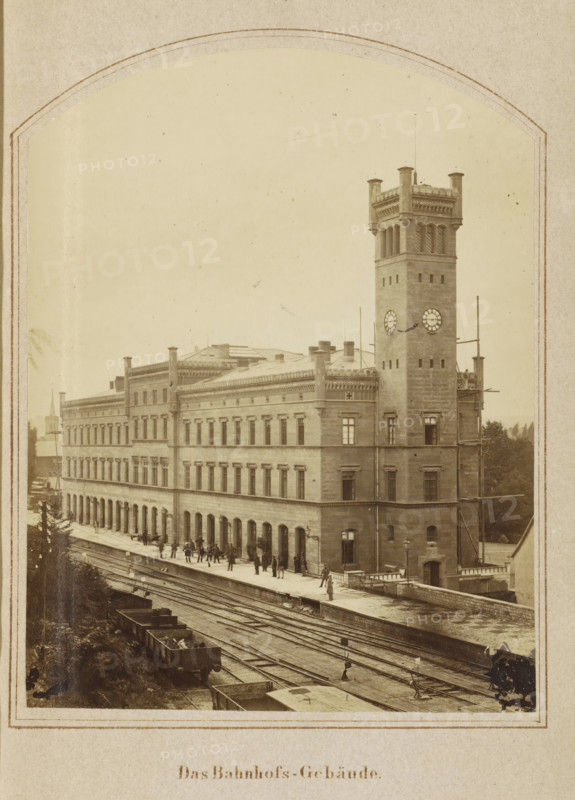 View of the Erfurt train station, Das Bahnhof-Gebäude, photograph, page, number: '8.', Erfurt (city), c. 1858 - in or before 1868, photographer, Thuringia, paper, albumen print, height 128 mm x width 98 mm, Image opposite page 100., station, shelter (railway, tramway), facade (of house or building), A sepia-toned photograph of a large stone building with rows of rectangular windows and decorative cornices, featuring a tall square clock tower with a crenellated top and round clock faces, a street-level platform beside multiple sets of railroad tracks with several open rail cars visible, small groups of people standing and walking along the platform, scaffolding attached to part of the tower, and trees visible in the background.