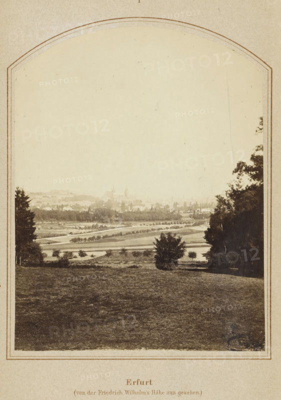 Erfurt in seiner Vergangenheit und Gegenwart, book, publisher: Carl Villaret, Erfurt (city), 1868, Thuringia, textile materials, pasteboard, paper, linen (material), printing, lithography, albumen print, height 185 mm x width 126 mm x thickness 14 mm, Book contains a fold-out map of Erfurt., A sepia-toned photograph framed by an arched border shows a broad grassy foreground leading down to a patchwork of fields and tree-lined paths, with scattered small trees and shrubs across the midground; a distant town with clustered buildings and a few taller spires rises faintly through haze on the horizon, and larger trees border the scene on the left and right edges.