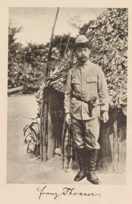 Portrait of Franz Thonner in front of a wooden house, Franz Thonner, page, photomechanical print, cliché maker: Stengel & Markert, Dresden, 1896, photographer, Saxony, paper, collotype, photogravure, height 162 mm x width 109 mm, Print after page X., Person, explorer, A sepia-toned photograph of a bearded man standing in front of a thatched structure with leafy covering, wearing a wide-brimmed hat, a buttoned jacket with flap pockets, a belt at the waist, trousers tucked into tall boots, and holding a long wooden staff; behind him are vertical poles and dense foliage, and a patch of ground and plants is visible at his feet.
