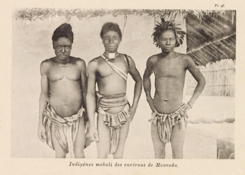 Portrait of three Mobali men near Monveda, Indigènes mobali des environs de Monveda, page, photomechanical print, number: 'Pl. 46.', photographer: Franz Thonner, Congo (country), cliché maker: Stengel & Markert, Dresden, 1896, Saxony, paper, collotype, photoprint, height 108 mm x width 162 mm, adult man, Three dark-skinned individuals stand side by side beneath a thatched roof, each wearing minimal cloth skirts and bare chests; the person on the left has a taller hair arrangement and a rounded abdomen, the central figure wears a strap across the chest and a small rectangular ornament at the neck, and the person on the right has short hair adorned with foliage and a hand resting on the hip, while a bamboo or wooden fence and a sloped thatched wall are visible in the background.