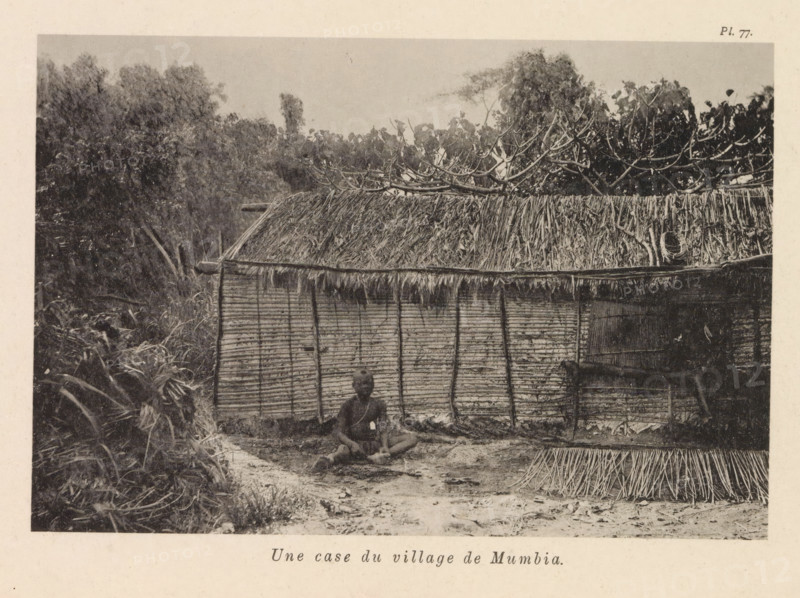 Exterior of a house in Mumbia, Une case du village de Mumbia, page, photomechanical print, number: 'Pl. 77.', photographer: Franz Thonner, Congo (country), cliché maker: Stengel & Markert, Dresden, 1896, Saxony, paper, collotype, light printing, height 108 mm x width 161 mm, adult man, housing, façade (of house or building), A sepia-toned photograph shows a thatched hut with a woven wall and a roof made of layered dried leaves, vegetation behind and above the roof including leafy branches, and a person sitting on the ground in front of the hut on a dirt area with scattered plant material.