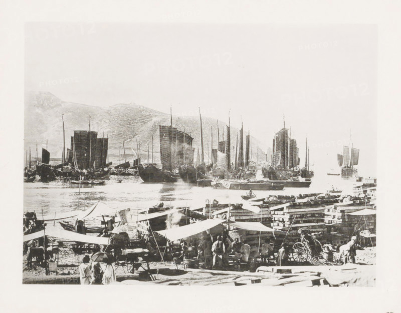 Drying place for sails in Port Arthur, with Chinese junks in the background, page, photomechanical print, number: '15', photographer: AN Ljupova, Port Arthur, cliché maker: anonymous, 1898 - 1899, Manchuria, paper, collotype, photoprint, height 150 mm x width 203 mm, ships (in general), sail, A busy waterfront scene with numerous traditional sailing vessels lined up on calm water, their tall masts and large rectangular sails prominent against a pale sky and distant hilly shoreline; closer to the viewer a crowded quay area shows market stalls and canvas awnings, stacks of crates and timber, and groups of people standing and moving about, some wearing wide-brimmed hats and carrying umbrellas, while small boats and rowboats dot the water near the larger ships.