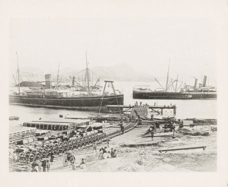 Jetty of the East China Railway at Port Arthur, page, photomechanical print, number: '17', photographer: AN Ljupova, Port Arthur, cliché maker: anonymous, 1898 - 1899, Manchuria, paper, collotype, photoprint, height 150 mm x width 203 mm, railway, tramway, rack railway, ships (in general), A harbor scene showing several large steamships moored in the water with tall masts and funnels, a wooden pier extending out with people standing and walking on it, a crane-like structure near the pier, rows of wagon wheels and stacked crates or beams arranged on the shore, horse-drawn carts and scattered figures of people working or resting on the beach, a track or wooden rails leading from the shore onto the pier, and distant low hills visible across the water.