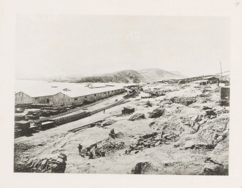 View of the East China Railway, page, photomechanical print, number: '19', photographer: AN Ljupova, Manchuria, cliché maker: anonymous, 1898 - 1899, paper, collotype, photoprint, height 150 mm x width 203 mm, warehouse (, harbour), railway, tramway, rack railway, A wide black-and-white scene showing a rocky, uneven shoreline with scattered stones and rough ground in the foreground, a few small groups of people working among tools and carts, a set of parallel railway tracks running along the midground beside long warehouse buildings with peaked roofs, stacks of materials and circular coils near the tracks, a curved dirt road leading toward a cluster of low buildings and structures further back, rolling hills rising behind the buildings, and a pale sky above.