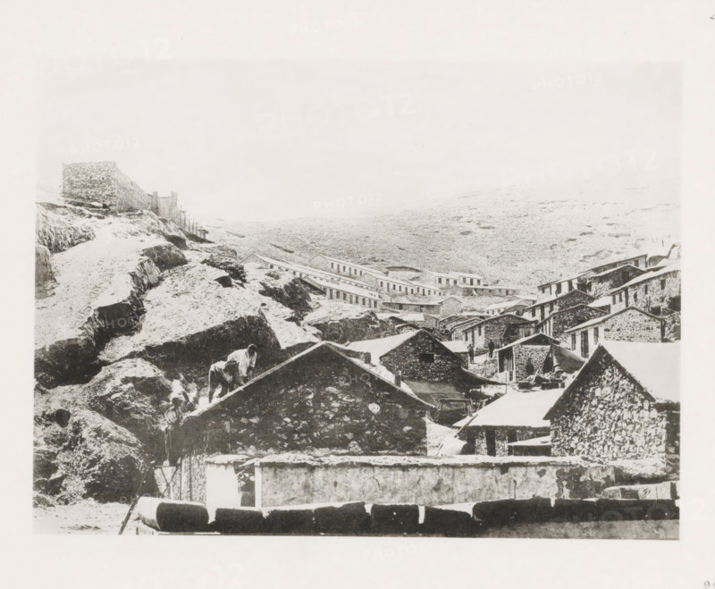 Workers' houses along the slope of a mountain in Port Arthur, page, photomechanical print, number: '23', photographer: AN Ljupova, Port Arthur, cliché maker: anonymous, 1898 - 1899, AN Ljupova, photographer, cliché maker, Manchuria, paper, collotype, light print, height 150 mm x width 203 mm, working class, labourers, (high) hill, housing, facade (of house or building), A sepia-toned scene shows a cluster of stone buildings with sloped roofs arranged on a rocky hillside, with terraces and retaining walls visible among rough outcrops. A steep rocky path winds up the left side toward a stone wall with a small structure, and a row of low long buildings stretches across the midground along the slope. In the foreground several pitched roofs and stone facades form a dense arrangement, with a few indistinct human figures near the buildings and someone on a roof. The landscape behind the settlement is a barren, gently sloping expanse with scattered rocks, and the sky above is pale and largely ...