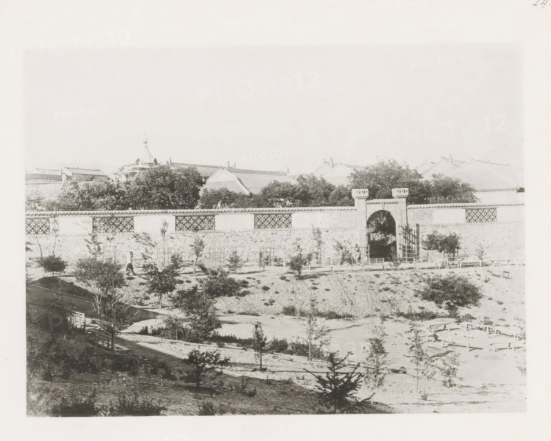 View of the Nicholas Boulevard in Port Arthur, page, photomechanical print, number: '24', photographer: AN Ljupova, Port Arthur, cliché maker: anonymous, 1898 - 1899, Manchuria, paper, collotype, light pressure, height 150 mm x width 203 mm, city-walls, avenue, boulevard in village, A wide view shows a long stone wall with decorative lattice sections and a central arched gateway topped by short battlement-like structures, behind which tree canopies rise and tiled roofs with ornate ridges are visible; in front of the wall there is an embankment with scattered small trees and shrubs, simple wooden fences and walkways, and a few people near the gateway, all rendered in a faded monochrome tone.