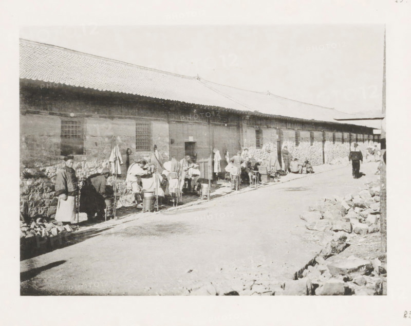 Street barbers in Port Arthur, page, photomechanical print, number: '25', photographer: AN Ljupova, Port Arthur, cliché maker: anonymous, 1898 - 1899, Manchuria, paper, collotype, photoprint, height 150 mm x width 203 mm, street, barber, barbershop, shaving, being shaved, A long single-story building with a tiled roof and stone lower walls extends along a street, with several rectangular windows and a central doorway; a line of people stand and sit along the building’s side, some wearing cloaks and hats and others with aprons, gathered around buckets, stools, and small stands; clothing and bundled items hang on hooks or lean against the wall; a man walks along the street away from the group toward the right side of the image; piles of stones and rubble are scattered along the curb in the foreground and near the right edge; shadows fall across the street and the overall scene appears to be in monochrome.