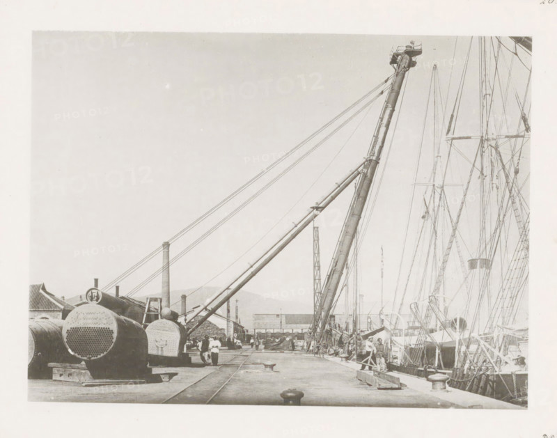 Port crane at Port Arthur, page, photomechanical print, number: '26', photographer: AN Ljupova, Port Arthur, cliché maker: anonymous, 1899 - 1898, Manchuria, paper, collotype, photoprint, height 150 mm x width 203 mm, harbour, dock crane, A dock scene showing large cylindrical metal objects with perforated ends stacked on the left, a long angled crane boom extending upward from the dock toward the right, multiple rigging lines and masts of ships rising along the right edge, workers and figures standing and walking along the pier, tracks embedded in the pier surface, bollards and wooden planks on the dock, industrial buildings and chimneys in the background, and distant hills beneath a pale sky.