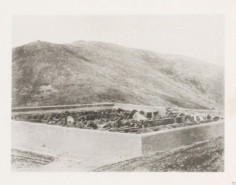 Burned Chinese camp near Port Arthur, page, photomechanical print, number: '28', photographer: AN Ljupova, Manchuria, cliché maker: anonymous, 1898 - 1899, paper, collotype, photoprint, height 150 mm x width 203 mm, ruin of a building, architecture, mountains, A grainy black-and-white photograph showing a low stone wall or rectangular enclosure in the foreground with rough, irregular stones and sections of smoother masonry, and behind it a large sloping hillside covered in textured, mottled terrain extending across the image under a pale sky.