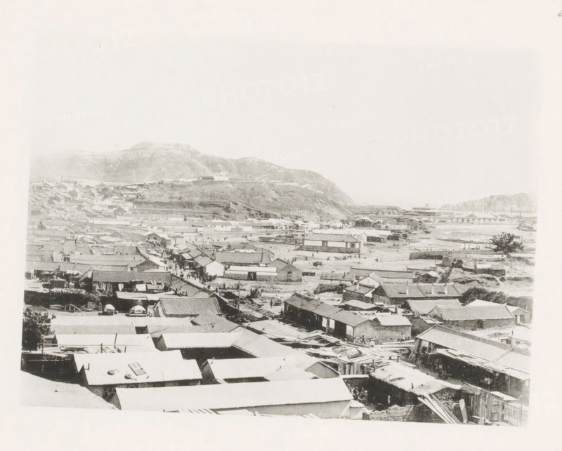 View of Port Arthur, page, photomechanical print, number: '6', photographer: AN Ljupova, Port Arthur, cliché maker: anonymous, 1898 - 1899, Manchuria, paper, collotype, photoprint, height 150 mm x width 203 mm, city-view in general, 'veduta', facade (of house or building), A wide grayscale view shows a densely packed town with many low buildings and tiled roofs extending toward a shoreline, with open sandy areas and scattered small boats near the water; a hillside rises behind the town with terraced slopes and a few isolated structures, and distant cliffs frame the right edge of the scene, while narrow streets and alleys run between rows of houses with occasional groups of small figures visible among them.