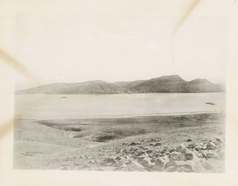 View of the bay at Port Arthur, page, photomechanical print, number: '9', photographer: AN Ljupova, Manchuria, cliché maker: anonymous, 1898, paper, collotype, photoprint, height 150 mm x width 203 mm, gulf, bay, sea, A wide, pale landscape photograph showing a calm body of water bordered by distant rolling hills and small mountains across the horizon, with a shoreline in the foreground that transitions from rocky terrain to flatter, grassy ground; two small boats float on the water near the right side and another small vessel is visible farther left; the sky above is light and largely empty, with subtle discoloration marks near the upper corners and faint variations in tone across the image.