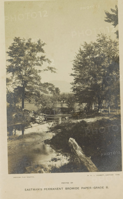 View of a ditch (presumably) in a park, photograph, page, photographer: WJ Hickmott, Connecticut, c. 1884 - c. 1889, North America, paper, gelatin silver print, height 164 mm x width 108 mm, public gardens, park, A sepia-toned landscape photograph showing a narrow stream flowing toward the foreground with reflective water and a fallen log lying along the near bank, trees with dense foliage flanking both sides of the stream, a simple wooden fence crossing the stream on a small bridge, grassy and brushy banks with scattered rocks, and distant low hills visible through the trees under a pale sky.
