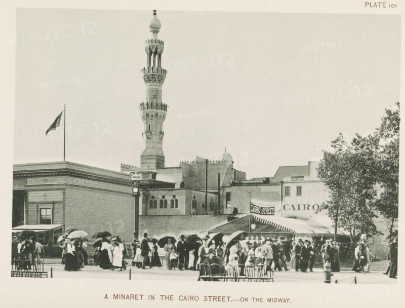 View of a replica of a street from Cairo with a minaret at the World's Columbian Exposition in Chicago in 1893, A minaret in the Cairo street.- on the Midway, page, photomechanical print, number: 'Plate 101', photographer: Charles Dudley Arnold, Chicago, photographer: HD Higinbotham, clichémaker: anonymous, 1893, Charles Dudley Arnold, HD Higinbotham, photographer, clichémaker, North America, Illinois, paper, collotype, photogravure, height 133 mm x width 191 mm, temple, shrine, Islam, Mohammedanism, world fair, world exhibition, World's Columbian Exposition, A city street scene with a tall ornate minaret rising above a row of flat-roofed buildings, a flag flying from a pole on one rooftop, a lamppost in front of a building with arched windows, a canopyed awning extending over part of the sidewalk, a large tree on the right, and many people wearing early twentieth-century clothing and carrying umbrellas or hats gathered and walking in the foreground.