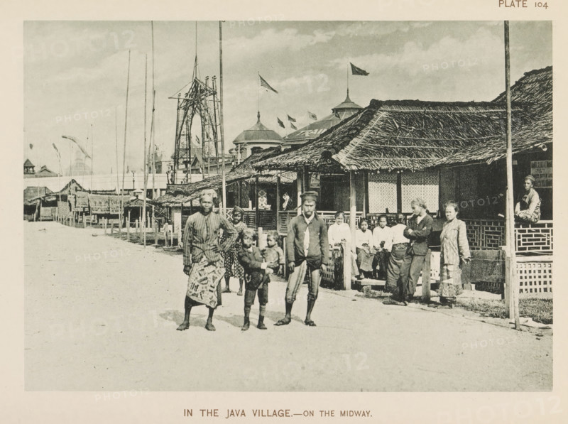 Houses and people from Java at the World's Columbian Exposition in Chicago in 1893, In the Java village.- on the Midway, page, photomechanical print, number: 'Plate 104', photographer: Charles Dudley Arnold, Chicago, photographer: HD Higinbotham, cliché maker: anonymous, 1893, Charles Dudley Arnold, HD Higinbotham, photographer, cliché maker, North America, Illinois, paper, collotype, photoprint, height 133 mm x width 190 mm, world fair, world exhibition, World's Columbian Exposition, A wide street scene shows a row of thatched-roof buildings with woven walls and open fronts along one side, with tall poles and flagpoles rising behind them and a pavilion-like structure with domed roofs visible further back; a group of people stands and sits near the buildings, including adults and children wearing patterned cloth wraps and shirts, some barefoot and some leaning on railings, while one person is seated on a raised platform; in the foreground a few figures walk toward the camera on a broad...