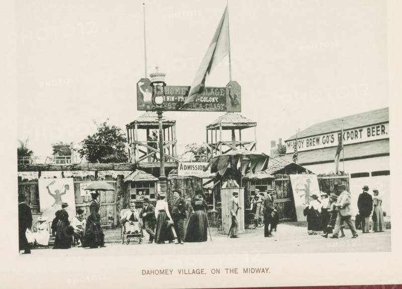 Replica of a Dahomey village at the World's Columbian Exposition in Chicago in 1893, Dahomey village, on the Midway, page, photomechanical print, number: 'Plate 110', photographer: Charles Dudley Arnold, Chicago, photographer: HD Higinbotham, cliché maker: anonymous, 1893, Charles Dudley Arnold, HD Higinbotham, photographer, cliché maker, North America, Illinois, paper, collotype, photoprint, height 132 mm x width 191 mm, world fair, world exhibition, World's Columbian Exposition, A crowded outdoor scene shows a wooden entrance structure with two raised watchtower-like platforms, a tall lamppost in front, and a large flag flying above one side; beneath the raised platforms are shaded booths with awnings and a sign hanging centrally; a group of people in period clothing stand and walk in the foreground, including women in long dresses and hats, men in suits and hats, and some seated figures and a person in a small wheeled chair; wooden fencing and painted display panels flank the entran...