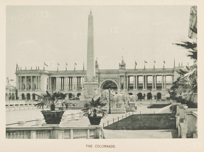 The colonnade, View of a colonnade and an obelisk at the World's Columbian Exposition in Chicago in 1893, page, photomechanical print, number: 'Plate 20', photographer: Charles Dudley Arnold, Chicago, photographer: HD Higinbotham, cliché maker: anonymous, 1893, Charles Dudley Arnold, HD Higinbotham, photographer, cliché maker, North America, Illinois, paper, collotype, photoprint, height 133 mm x width 191 mm, colonnade, obelisk, needle, world fair, world exhibition, World's Columbian Exposition, A wide plaza with a tall obelisk at its center stands in front of a large neoclassical building with a long colonnade and decorative balustrade; flagpoles line the roof and sculptural figures sit atop sections of the structure. In front of the obelisk is a raised sculptural group including a pair of large animal figures and ornate pedestals, flanked by planters containing palm-like plants. A formal path and low stone fencing run across the foreground, with trimmed grassy areas bordered by shor...