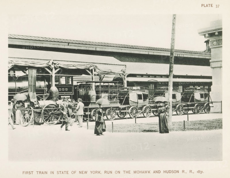 First train in State of New York, run On The Mohawk & Hudson R., R., 1831, First train to run on the Mohawk & Hudson Railroad, exhibited at the World's Columbian Exposition in Chicago in 1893, page, photomechanical print, number: 'Plate 37', photographer: Charles Dudley Arnold, Chicago, photographer: HD Higinbotham, clichémaker: anonymous, 1893, Charles Dudley Arnold, HD Higinbotham, photographer, clichémaker, North America, Illinois, paper, collotype, photoprint, height 131 mm x width 191 mm, railway, train, world fair, world exhibition, World's Columbian Exposition, A sepia-toned scene shows a train platform with a steam locomotive and a row of passenger carriages under a canopy with draped fabric, large spoked wheels visible beneath the vehicles, and a vertical pole in the foreground; several people in period clothing walk or stand nearby, some holding umbrellas, while architectural details of the station and decorative stonework appear at the right edge.