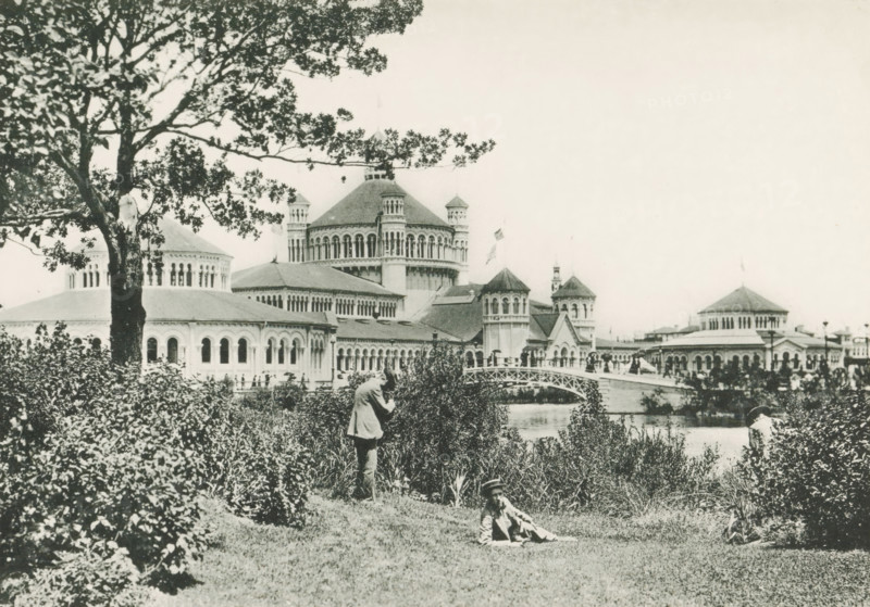 Fisheries Builing from Wooded Island, View of the fishery building, designed by Henry Ives Cobb at the World's Columbian Exposition in Chicago in 1893, page, photomechanical print, number: 'Plate 53', photographer: Charles Dudley Arnold, Chicago, photographer: HD Higinbotham, cliché maker: anonymous, 1893, Charles Dudley Arnold, HD Higinbotham, photographer, cliché maker, North America, Illinois, paper, collotype, photoprint, height 132 mm x width 190 mm, world fair, world exhibition, World's Columbian Exposition, A black and white photograph showing a large ornate building complex with domes, arched windows, and towered structures along a waterfront, partially obscured by a large tree and dense shrubbery in the foreground; a curved pedestrian bridge spans the water near the center, and people are visible on the bridge and along the shoreline; in the grassy foreground a person stands among the bushes while another person reclines on the grass wearing a hat; the sky appears light and em...