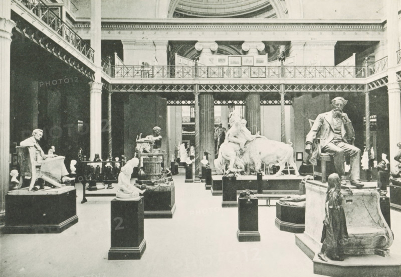 Interior of a room with sculptures in the Palace of Fine Arts at the World's Columbian Exposition in Chicago in 1893, Interior of Palace of Fine Arts, page, photomechanical print, number: 'Plate 61', photographer: Charles Dudley Arnold, Chicago, photographer: HD Higinbotham, cliché maker: anonymous, 1893, Charles Dudley Arnold, HD Higinbotham, photographer, clichémaker, North America, Illinois, paper, collotype, photoprint, height 134 mm x width 191 mm, piece of sculpture, reproduction of a piece of sculpture, permanent exhibition, museum, world fair, world exhibition, World's Columbian Exposition, Palace of Fine Arts, A large indoor gallery with high ceilings and a two-tiered iron and columned balcony running along the upper level, filled with numerous sculptures on pedestals and plinths arranged across the floor; visible statues include seated figures, a reclining animal with figures on its back, busts, and a standing figure in the foreground on a raised base.