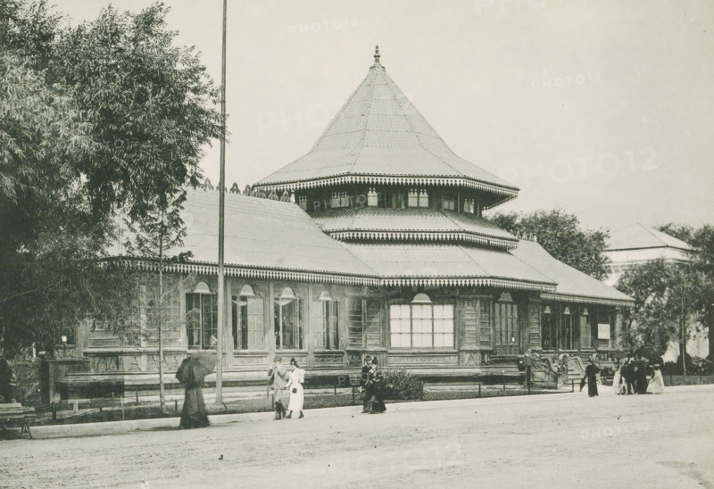 Pavilion of Ceylon at the World's Columbian Exposition in Chicago in 1893, The Ceylon Building, page, photomechanical print, number: 'Plate 79', photographer: Charles Dudley Arnold, Chicago, photographer: HD Higinbotham, cliché maker: anonymous, 1893, Charles Dudley Arnold, HD Higinbotham, photographer, cliché maker, North America, Illinois, paper, collotype, photoprint, height 134 mm x width 190 mm, world fair, world exhibition, World's Columbian Exposition, A large wooden pavilion with a wide, multi-tiered roof and decorative eaves occupies the center of the image, flanked by trees on either side and a tall slender pole in front; the building shows multiple tall windows and ornate carved details along its base and cornices, with a conical central roof topped by a finial. In front of the pavilion a few people in period clothing stand and walk along a paved area, some holding umbrellas and others gathered in small groups, while low fencing and a bench are visible near the building and ...