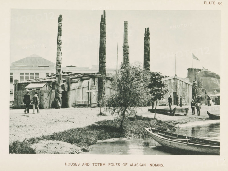 Houses and totem poles of Alaskan people at the World's Columbian Exposition in Chicago in 1893, Birchbark wigwams of Penobscot Indians, page, photomechanical print, number: 'Plate 89', photographer: Charles Dudley Arnold, Chicago, photographer: HD Higinbotham, cliché maker: anonymous, 1893, Charles Dudley Arnold, HD Higinbotham, photographer, cliché maker, North America, Illinois, paper, collotype, photoprint, height 131 mm x width 191 mm, world fair, world exhibition, World's Columbian Exposition, A waterfront scene shows a row of tall carved wooden poles rising behind a cluster of wooden buildings with plank walls and pitched roofs, a small tree and a larger bush in front of the buildings, a wooden canoe moored at the water's edge, several men in period clothing walking or standing near the buildings and along the shore, a low dock or bench area by the water, and a distant hillside with a flag on a pole.