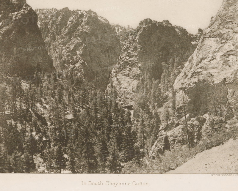 In South Cheyenne Cañon, View of the mountains near South Cheyenne Canyon, page, photomechanical print, photographer: WH Jackson Photograph and Publishing Co., Colorado Springs, cliché maker: New York Photo-Gravure Company, New York (city), c. 1888 - in or before 1893, North America, Colorado, paper, photoaquatint, height 161 mm x width 201 mm, Print opposite title page., landscapes, mountains, A sepia-toned photograph of a rugged canyon scene showing steep rocky cliffs rising from a densely forested valley, with tall evergreen trees filling the lower areas and clinging to ledges, rough textured rock faces with visible strata and crevices, a sloping foreground of lighter rocky ground at the bottom right, and a pale sky above.