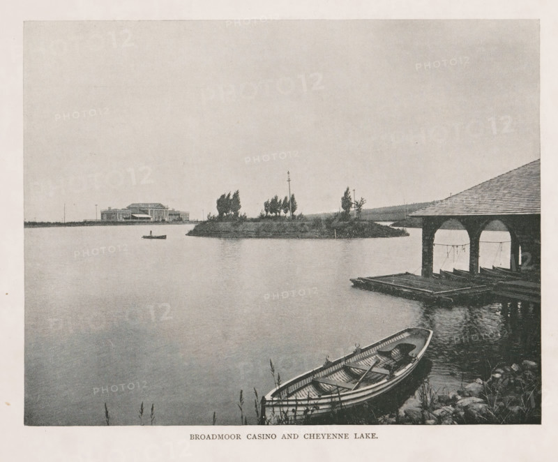 View of the Broadmoor Casino and Cheyenne Lake in Colorado Springs, Broadmoor Casino and Cheyenne Lake, page, photomechanical print, Colorado Springs, cliché maker: New York Photo-Gravure Company, New York (city), c. 1888 - in or before 1893, photographer, North America, Colorado, paper, autotypie, height 157 mm x width 209 mm, lake, casino, A wide view of a calm lake with a small island topped by trees and a tall slender pole near its center, a distant building with a low roofline visible on the far shore, a lone rowboat floating on the water near the horizon, a covered boathouse with open arched bays extending over the water at the right edge, wooden docks beneath the boathouse with more boats stored inside, a foreground shoreline showing a moored rowboat with visible oars and interior seats, and rippling reflections on the water surface.