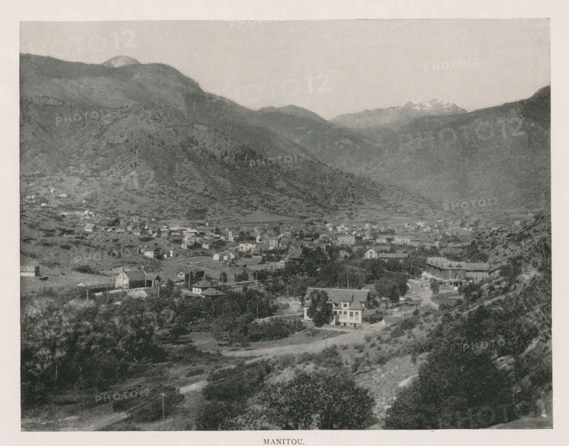 Manitou, View of Manitou Springs, page, photomechanical print, Manitou Springs, cliché maker: New York Photo-Gravure Company, New York (city), c. 1888 - in or before 1893, photographer, North America, Colorado, El Paso (Colorado), paper, autotypie, height 154 mm x width 201 mm, city-view in general, 'veduta', mountains, A wide landscape photograph shows a valley town nestled among rolling hills and steep mountains, with numerous small buildings and houses spread across the flat area and lower slopes, a few larger structures standing out, tree clusters and open grassy areas interspersed between the buildings, a road winding through the foreground past houses and vegetation, rocky hillsides and scattered shrubs rising toward the base of larger, darker mountain ridges that dominate the middle ground, and distant higher peaks visible along the horizon beneath a pale sky.