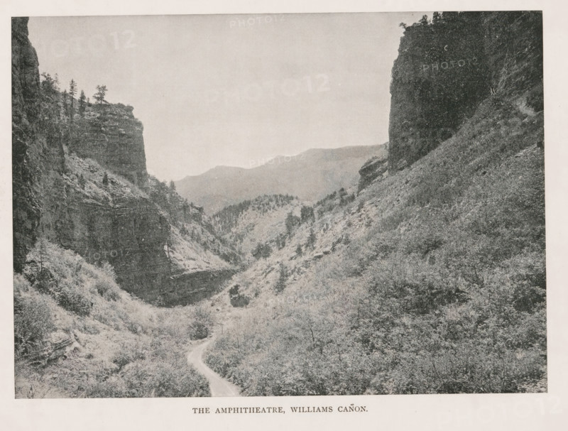 The Amphitheatre, Williams Cañon, View of the Williams Canyon, page, photomechanical print, Williams Canyon (El Paso, CO), cliché maker: New York Photo-Gravure Company, New York (city), c. 1888 - in or before 1893, photographer, North America, Colorado, El Paso (Colorado), paper, autotypie, height 146 mm x width 203 mm, landscapes, ravine, chasm, abyss, canyon, A high-contrast black-and-white landscape photograph showing a narrow winding dirt road leading into a deep canyon flanked by steep rocky cliffs and densely textured vegetation. The left side of the image features a tall cliff face with layered rock strata and a few sparse trees along its top edge, while the right side shows a darker, sheer rock wall descending to a slope covered in shrubs and underbrush. In the middle distance the canyon opens to reveal rolling ridges and tree-covered hills fading into a lighter-toned sky that occupies the upper portion of the image.
