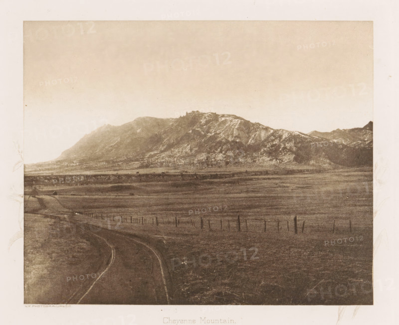 View of Cheyenne Mountain, Cheyenne Mountain, page, photomechanical print, photographer: WH Jackson Photograph and Publishing Co., Colorado, cliché maker: New York Photo-Gravure Company, New York (city), c. 1888 - in or before 1893, North America, paper, photoaquatint, height 144 mm x width 177 mm, landscapes, mountains, El Paso (Colorado), A sepia-toned landscape showing a broad plain in the foreground with a dirt road curving away toward the horizon, a line of posts and wire fences running parallel across the field, and a distant mountain range with rugged peaks and ridges under a pale sky.