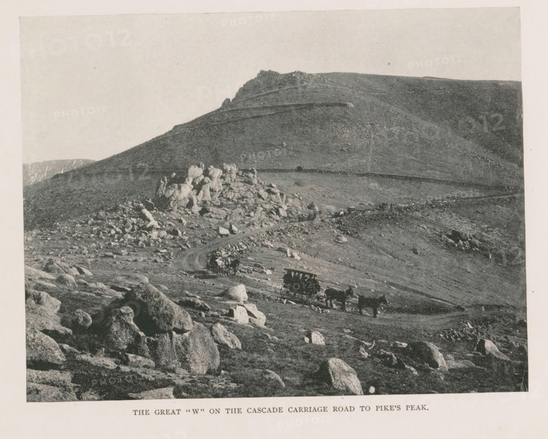 View of Pikes Peak and two carriages, The great W on the the cascade carriage road to Pike's Peak, page, photomechanical print, Colorado, cliché maker: New York Photo-Gravure Company, New York (city), c. 1888 - in or before 1893, New York Photo-Gravure Company, photographer, cliché maker, North America, paper, autotypie, height 154 mm x width 201 mm, four-wheeled vehicle drawn by more than three animals, mountain-top, peak, El Paso (Colorado), Pikes Peak (El Paso), A grayscale landscape showing a rocky hillside and ridge with scattered boulders and outcrops, a winding dirt road cutting across the slope, several horse-drawn wagons and teams traveling along the road, and a distant rocky peak and plateau under a pale sky.