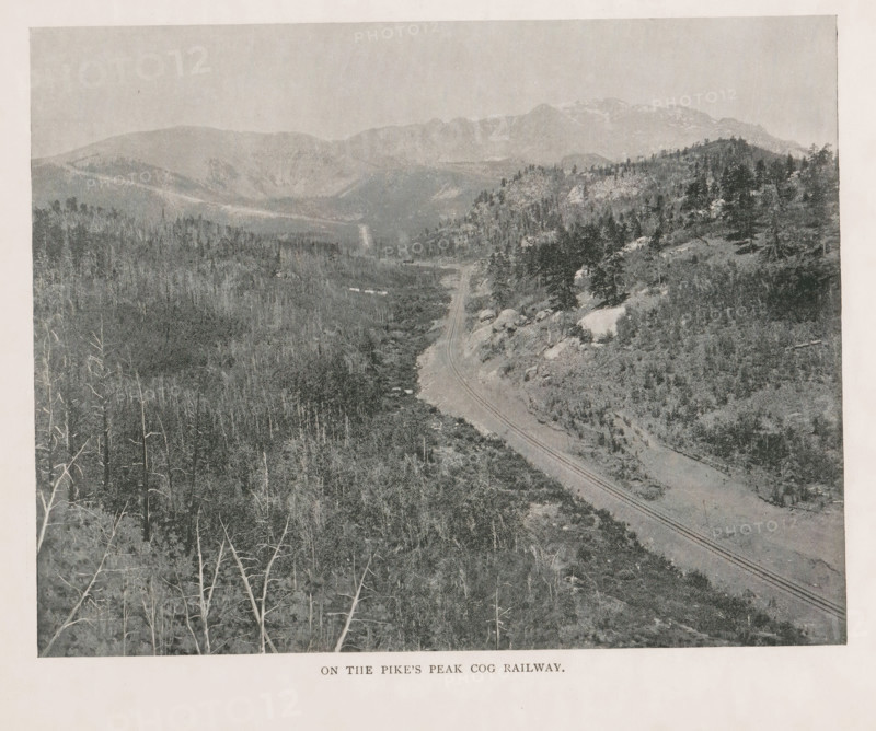 View of a cloud cover from Pikes Peak, Above the clouds, summit of Pike's Peak, page, photomechanical print, Colorado, cliché maker: New York Photo-Gravure Company, New York (city), c. 1888 - in or before 1893, New York Photo-Gravure Company, photographer, cliché maker, North America, paper, autotypie, height 90 mm x width 163 mm, clouds, mountain-top, peak, El Paso (Colorado), Pikes Peak (El Paso), A grayscale landscape photograph showing a winding railway track running through a valley with rocky slopes and scattered trees, a mix of bare trunks in the foreground and denser coniferous stands on the hillsides, with distant layered mountain ridges and a pale sky above.
