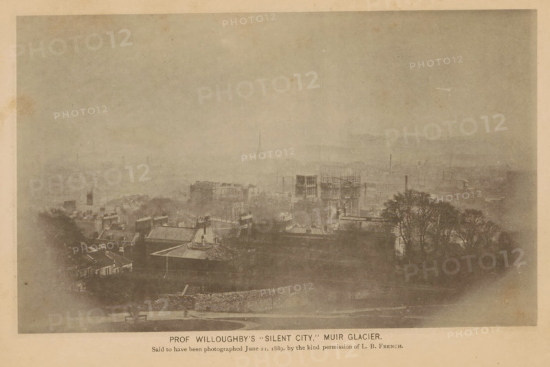View of the imaginary city of Silent City in Alaska, Prof. Willoughby's Silent City, Muir Glacier, page, photomechanical print, inscription: 'Said to be photographed June 21, 1889, by the kind permission of LB French.', c. 1885 - in or before 1890, paper, collotype, photoprint, height 113 mm x width 184 mm, city-view in general, 'veduta', North America, Alaska, Skagway-Hoonah-Angoon, A faded, sepia-toned view of a cityscape seen from an elevated vantage, with rooftops and building facades in the foreground, several taller structures and a scaffolding-covered building toward the middle ground, and distant hazy buildings and a barely visible spire receding into a pale sky, with a cluster of leafed trees on the right and a low stone wall and a bench near the bottom edge.