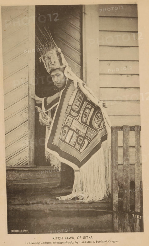 Portrait of Kitch Kawk of the Tlingit people, Kitch Kwak, of Sitka, page, photomechanical print, inscription: 'In Dancing Costume, photograph 7983, by Partridge, william henry, Portland, Oregon.', number: '7983', photographer: William Henry Partridge, Sitka, cliché maker: Britton & Rey, San Francisco, c. 1885 - in or before 1890, North America, California, Alaska, paper, collotype, photoprint, height 183 mm x width 109 mm, Person, Kitch Kawk, historical persons, city-view in general, 'veduta', glacier, A person stands in a doorway wearing a tall headdress with long upright feathers and a decorative band, a cloth draped over one shoulder, and a fringed rectangular garment with bold geometric patterns covering the front and extending toward the ground; the person has a ring in their nose and is holding the doorframe with one hand while the other hand grasps a carved object, and the scene shows wooden siding and planks around the doorway and beneath the person’s feet.